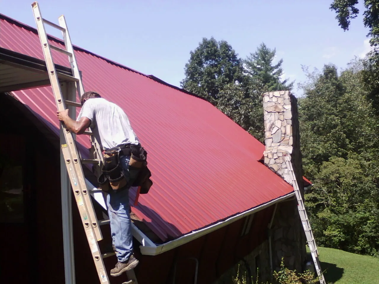 Roofer on a ladder beside a red metal roof and stone chimney — Soffit & Fascia Repair by Roofing Brothers in Palouse